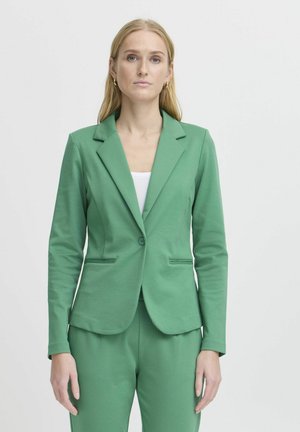 Vrouw met lang blond haar, goudkleurige oorringen en een groen colbert en broek over een wit shirt, staand tegen een effen achtergrond.