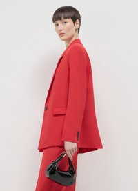 Blazer e pantaloni rossi in tessuto liscio, con una vestibilità sartoriale, maniche ampie e una borsa nera lucida tenuta in mano.