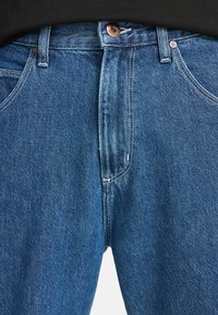 Niebieskie dżinsy o wysokim pasie, z miedziowanym guzikiem i standardowym pięciodotykowym stylem z widocznymi szwami.
