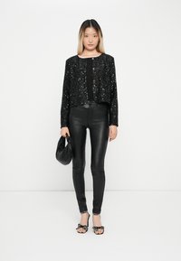 Fekete flitteres cropped dzseki egy testhezálló fekete top felett, fekete bőrhatású nadrággal párosítva, és nyitott orrú fekete magassarkú szandálokkal, masnival.