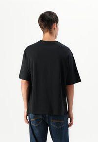 Egy férfi sima fekete, oversized pólót és kék farmert visel, háttal áll a kamerának, fehér háttér előtt.