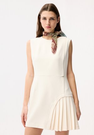 Frau trägt ärmelloses weißes Kleid mit plissiertem Rockteil und floralem Schal, der um den Hals gebunden ist, und steht vor einem einfarbigen Hintergrund.