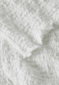 Tissu blanc texturé avec une surface froissée, présentant des bords doux et effilochés ainsi qu'un motif ondulé, mettant en valeur sa légèreté et sa qualité aérienne.