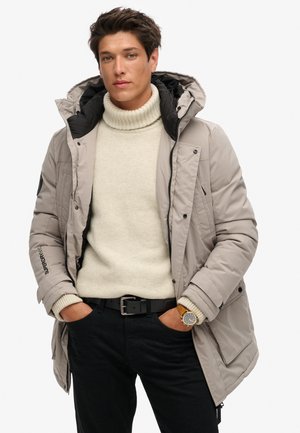 Chaqueta aislante gris claro con capucha, con bolsillos con cremallera y detalles en negro. Llevada sobre un suéter de cuello alto crema y pantalones negros.