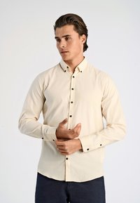Crème kleurig overhemd met lange mouwen en een button-down kraag. Heeft zwarte knopen aan de voorkant en op de manchetten; zachte textuur en een getailleerde pasvorm.