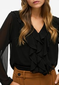 Blusa nera con dettagli arricciati sul davanti e maniche lunghe trasparenti, scollatura a V, abbinata a pantaloni marroni sartoriali con dettagli a bottone.