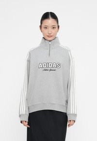 Mulher vestindo um moletom cinza Adidas com zíper de quarto e riscas brancas nas mangas, usando uma saia preta, de pé contra um fundo branco liso.