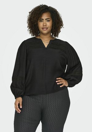 Blouse noire avec un col en V, tissu texturé et manches longues bouffantes. Dispose de coutures décoratives sur les épaules. Associée à un pantalon à motifs.