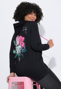 Zwarte hoodie met een grafisch ontwerp van een schedel en roze bloemen, textuurmateriaal en een roze koord. Ontspannen pasvorm en lange mouwen.