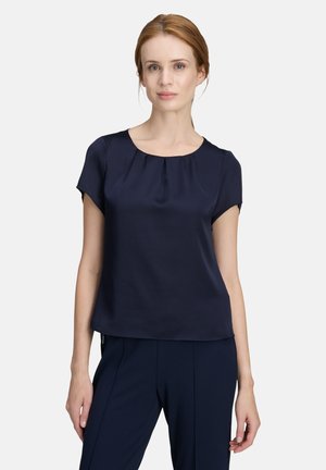 Woman with red hair wearing a navy blue short-sleeve blouse and matching navy pants, standing against a plain light background.