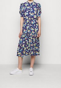 Květinové midi šaty v námořnické modři s krátkými nabíranými rukávy, zapínáním na knoflíky vpředu a volánkovým lemem. Vytvořeno s bílými teniskami s zlatými akcenty.