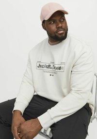 Crème sweatshirt met zwarte grafische tekst en patronen, met geribbelde manchetten, gecombineerd met een lichtroze pet. Zittende pose op een metalen stoel.