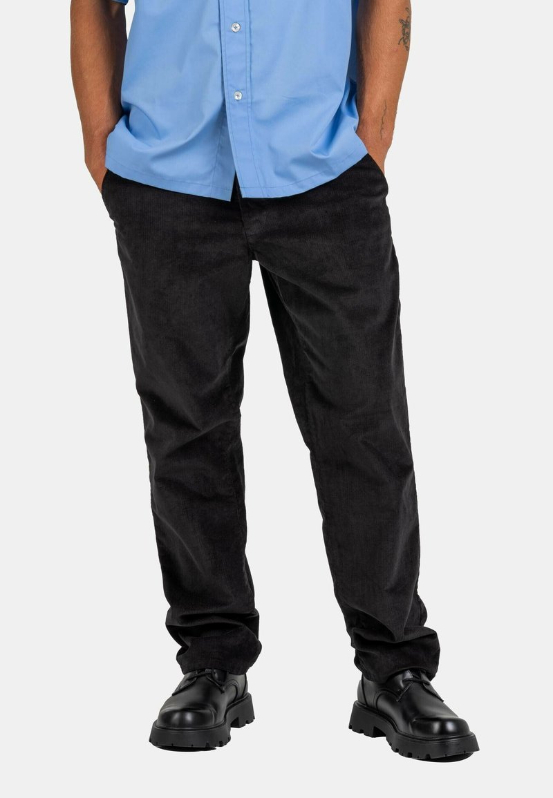 Black corduroy pants with a relaxed fit, featuring side pockets and a soft texture, paired with chunky black shoes and a light blue shirt.