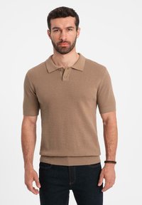 Brun kortärmad polo med texturerad väv, tvåknappsslå, ribbade ärmslut och figursydd midja, kombinerad med mörka jeans.