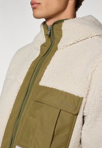 Soepe fleece-jakk, millel on oliivroheline vooder ja tõmblukk, millel on kreemikas värvi tekstuuriga väline osa ja esiküljel tasku logoga detailideks.