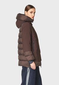 Chaqueta de plumas marrón con capucha, que presenta secciones acolchadas, bolsillos con cremallera y un acabado suave. Lleva una pantalón oscuro de campana.