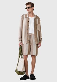 Beige und braun gestreifte Button-Up-Hemd, weißes, geripptes T-Shirt, passende Shorts, schwarze Slipper und eine grün gemusterte Tragetasche.