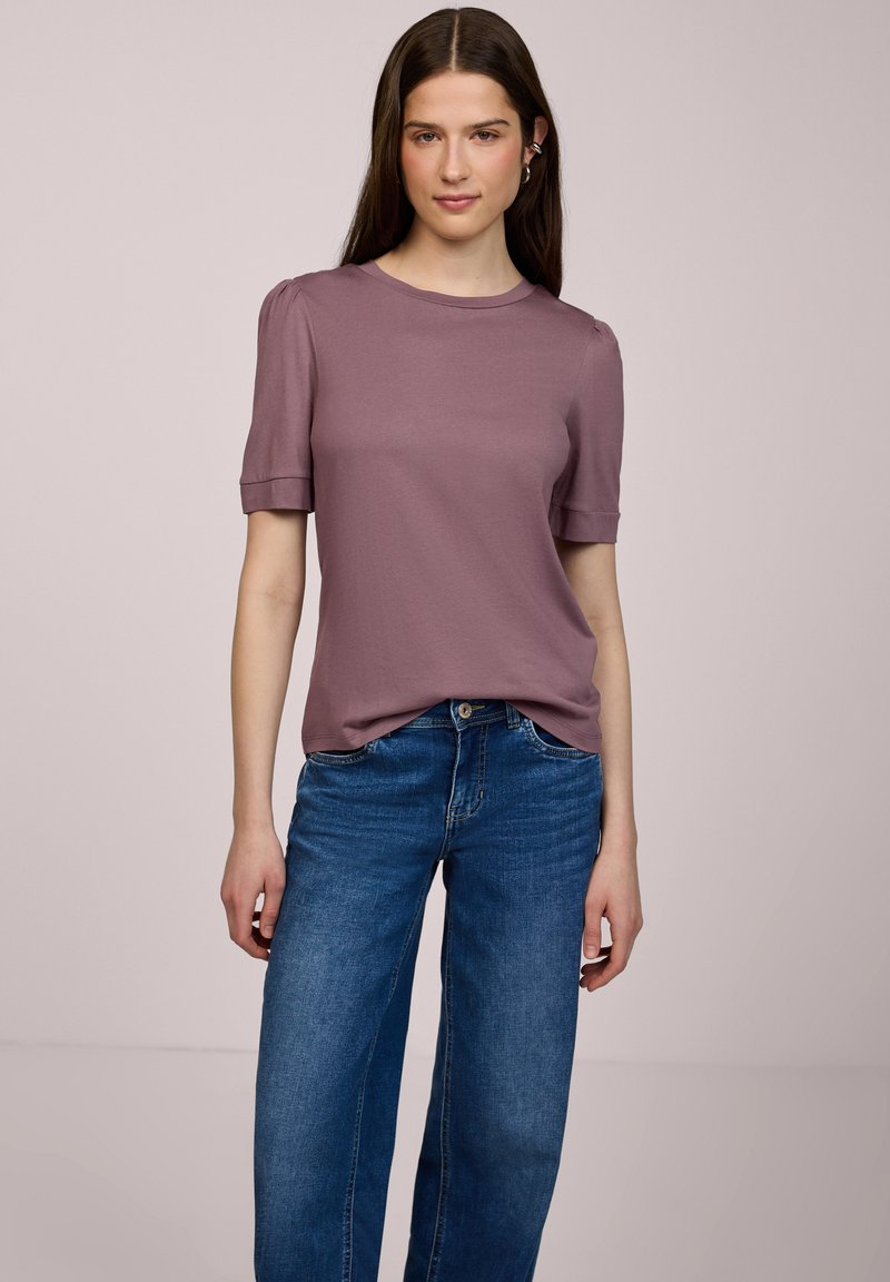 Ung kvinde står iført en mauve kortærmet top og blå højtaljede jeans mod en ensartet lys baggrund.