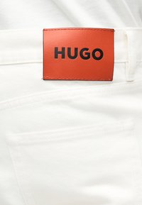 Λευκό τζιν από τζιν ύφασμα με πορτοκαλί ορθογώνιο δερμάτινο μπαλώμα ραμμένο πάνω από την πίσω τσέπη, με μαύρο κείμενο "HUGO".