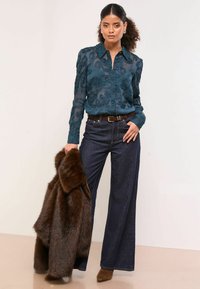 Priesvitná tyrkysová blúzka s kvetinovou textúrou, tmavé džínsy s širokými nohavicami, hnedý opasok a semišové čižmy po členky, nesúca hnedý kožuch.