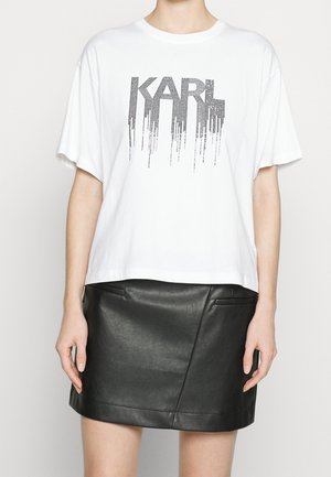 Persoon draagt een witte oversized t-shirt met de tekst "KARL" en een zwarte leren minirok, staat met armen ontspannen langs het lichaam.