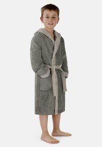 Grauer Kapuzenbadmantel mit einem weichen beigen Futter, Gürtel und zwei Fronttaschen. Hergestellt aus einem strukturierten, plüschigen Stoff. Das Modell steht barfuß.
