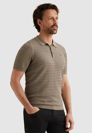 Mann mit Bart, der ein strukturiertes beiges kurzärmeliges Poloshirt trägt, steht mit einer Hand in der Tasche und schaut seitlich vor einem einfarbigen Hintergrund.