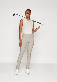 Beige ärmlös pikétröja med en texturerad mittdel, matchad med beiga tajta byxor. En golfklubba hålls i höger hand.