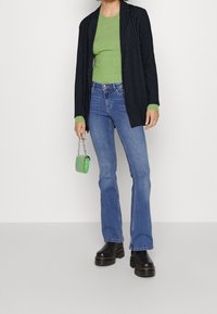 Person som bär en svart randig blazer, grön ribbad topp, blåjeans och svarta klumpiga stövlar, håller en liten grön handväska med kedja.