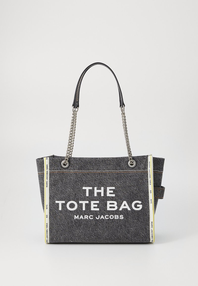 Sac fourre-tout en tissu gris avec des poignées en cuir noir et des accents en chaîne argentée. Présente un texte blanc indiquant "THE TOTE BAG MARC JACOBS" sur le devant.