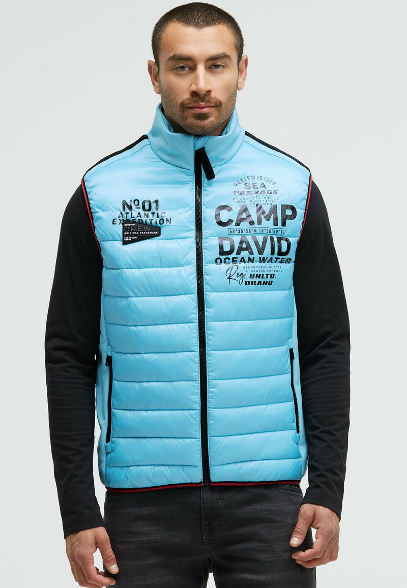 Hellblau gepolsterte Weste mit schwarzen Akzenten, die über einen hohen Kragen, Frontreißverschlüsse und bedruckte Texte verfügt, die die Marke und Designmerkmale hervorheben.