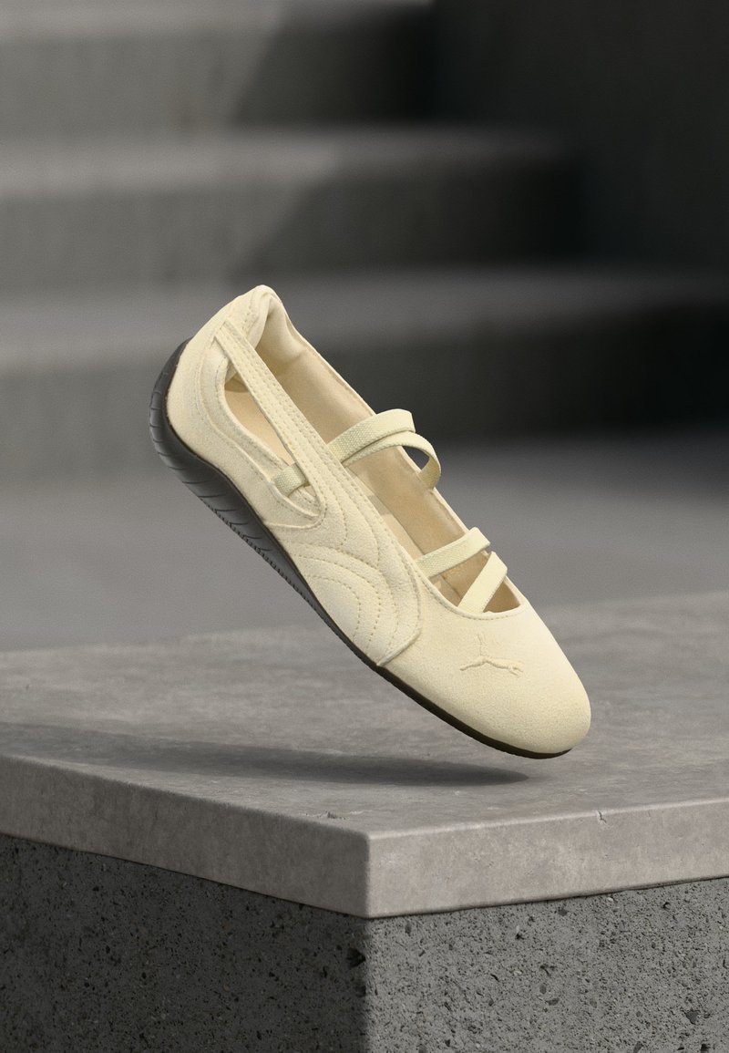 Ballerine couleur crème avec semelle noire et brides élastiques exposée sur un socle en béton gris avec un fond flou d'escaliers.