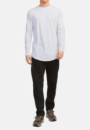 Camicia bianca a maniche lunghe con scollatura rotonda, abbinata a pantaloni neri e sneakers grigio-marroni; presenta una vestibilità rilassata e un design minimal.