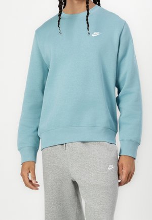 Personne portant un sweat-shirt Nike bleu clair et un pantalon de survêtement Nike gris, debout avec les bras détendus sur les côtés.