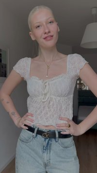 Ženska s svetlimi lasmi, oblečena v belo čipkasto kratko majico in svetlo modre kavbojke, stoji v notranjosti s postavljenimi rokami na bokih, nosi črni pas in zlato ogrlico.