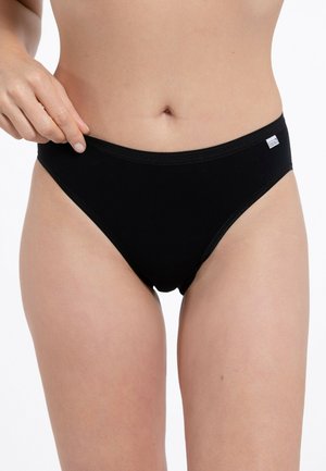 Donna che aggiusta mutandine nere in stile slip, mostrando la zona addominale e la parte superiore delle cosce su sfondo bianco.