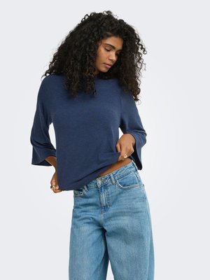 Femme aux cheveux bouclés portant un haut bleu à manches longues et un jean bleu clair taille haute, ajustant sa chemise sur un fond uni.