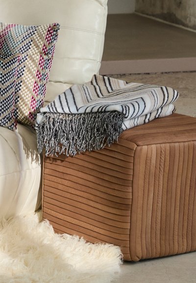 Ruskea raidallinen poufi, jolla on sileä pinta, kera taiteltu peitto, jossa on musta-valkoisia raitoja ja harmaa fringes.