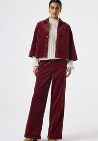 Bordeaux corduroy jasje en bijpassende wijde pantalon, aangevuld met een crèmekleurige kabelgebreide trui. Bevat metalen knopen en een zak.