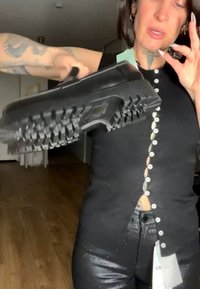 Persona che tiene una scarpa nera con suola spessa vicino alla fotocamera, indossa una maglia nera con bottoni e pantaloni neri lucidi all’interno.