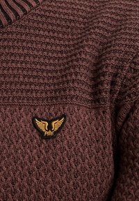 Brązowy sweter z dzianiny o teksturowanym wzorze. Posiada prążkowany dekolt oraz czarne haftowane logo z skrzydłami na obszarze klatki piersiowej.
