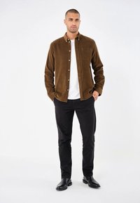 Cămașă din corduroy maro cu guler button-down, având un buzunar pe piept, purtată peste un tricou alb, asociată cu pantaloni negri și cizme.