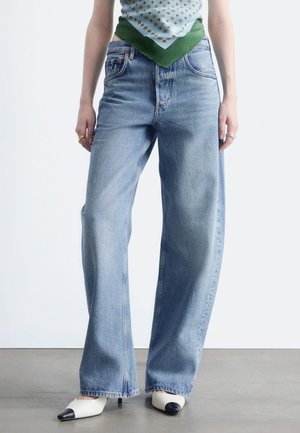 Persona che indossa jeans azzurri a gamba larga, un top bandana con motivi verdi e blu e scarpe bianche con punte nere, in piedi su pavimento grigio.