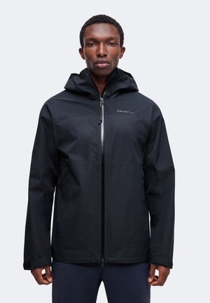 Homme portant une veste à capuche noire zippée, debout, mains détendues le long du corps, devant un fond clair uni.