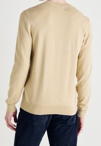 Man in een beige lange mouw sweater en donkerblauwe corduroy broek, getoond vanachter tegen een neutrale achtergrond.