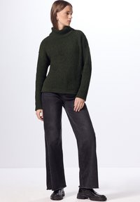 Maglione a coste verde scuro con colletto alto, abbinato a jeans neri larghi e stivaletti neri alla caviglia. La texture è corposa e calda.