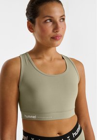 Sportowy biustonosz w jasnym oliwkowym zielonym kolorze z szerokimi ramionami, okrągłym dekoltem i dopasowanym krojem. Na przednim pasie znajduje się biały detal w postaci logo.