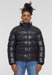 Zwarte pufferjack met een hoge kraag, glanzende afwerking, gestikte design en een ritssluiting aan de voorkant. Aangevuld met een lichte binnenvoering.