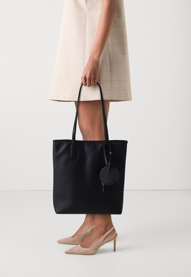 Femme en robe beige et talons tenant un grand sac cabas en cuir noir avec une petite pochette ronde attachée.