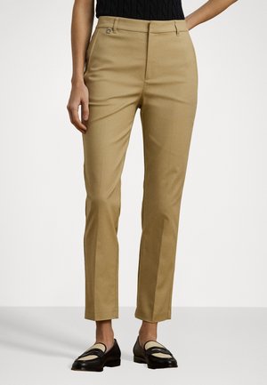 Vrouw draagt beige rechte broek gecombineerd met zwarte en beige loafers, staand tegen een effen lichte achtergrond.